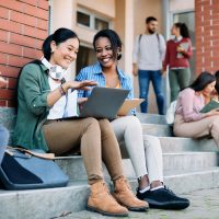 Happy multi-ethnic female students have fun while using laptop in front of university building.